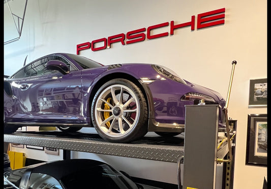 A Porsche dealership sign with non-LED red letters mounted on a wall, with a purple Porsche car displayed in front.
